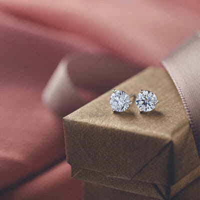 A pair of diamond stud earrings rests on a closed, gold-colored gift box with a pink fabric background and ribbon.