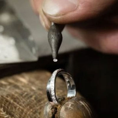 A hand sets a small round gemstone into a silver ring using a jewelry tool, with the ring held steady on a work surface.