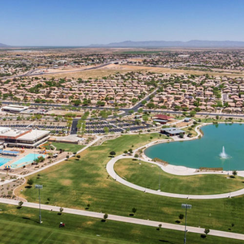Aerial view of a suburban neighborhood with houses, a large park, a pond with a fountain, sports fields, and swimming pools under a clear sky.
