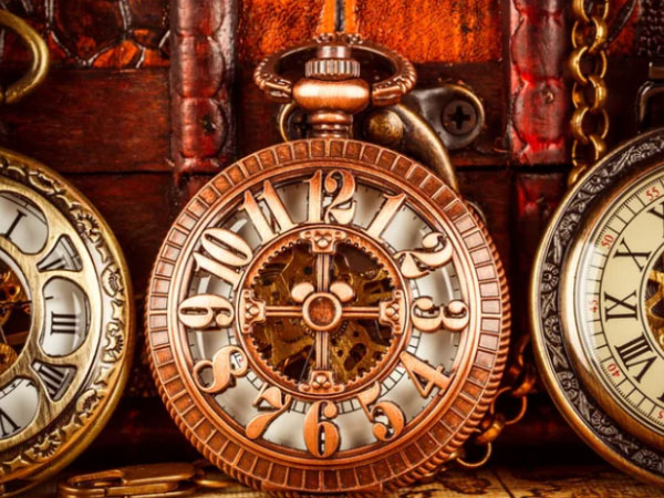 Three ornate antique pocket watches with visible gears are displayed against a wooden background. The central watch is bronze-colored and prominently featured in the foreground.