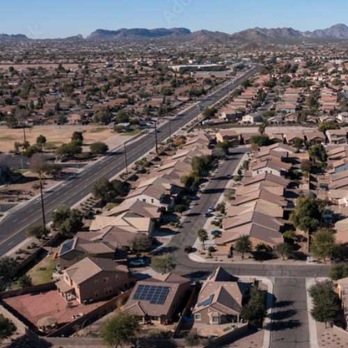 Aerial view of a suburban neighborhood with rows of houses, a main road, sparse trees, and mountains in the background under a clear sky.