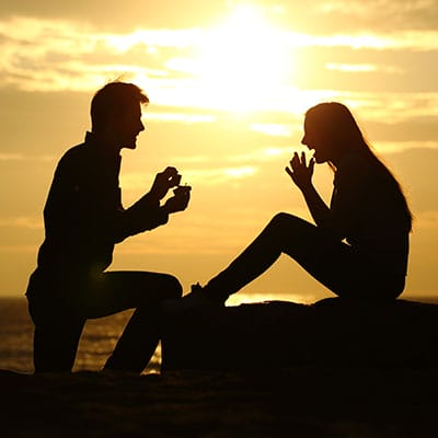 A person kneels and holds a ring box toward another person at sunset, silhouetted against the sky, suggesting a marriage proposal.