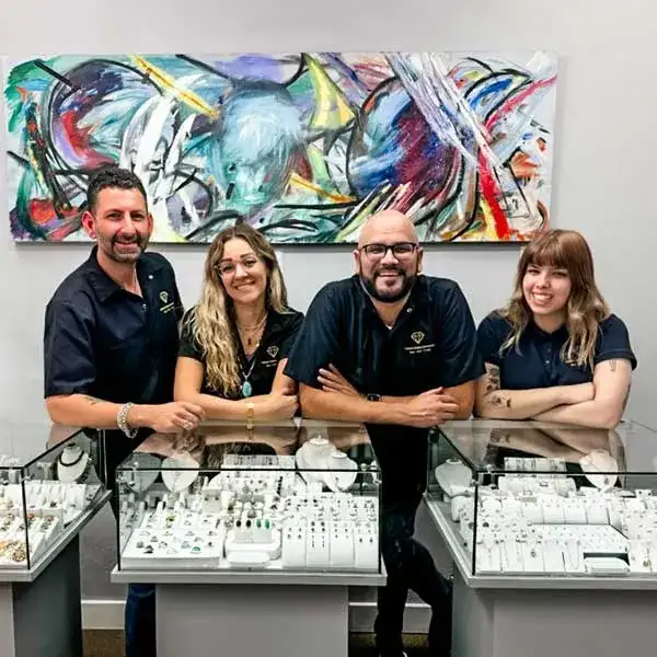 Four people stand behind two glass jewelry display cases, smiling, with a colorful abstract painting on the wall behind them.