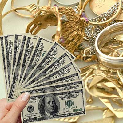 A hand holds several $100 bills in front of a pile of assorted gold jewelry, including bracelets, necklaces, and rings.