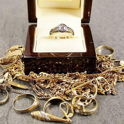 A silver ring with a clear stone in a box is displayed among several gold rings, necklaces, and bracelets on a gray surface.