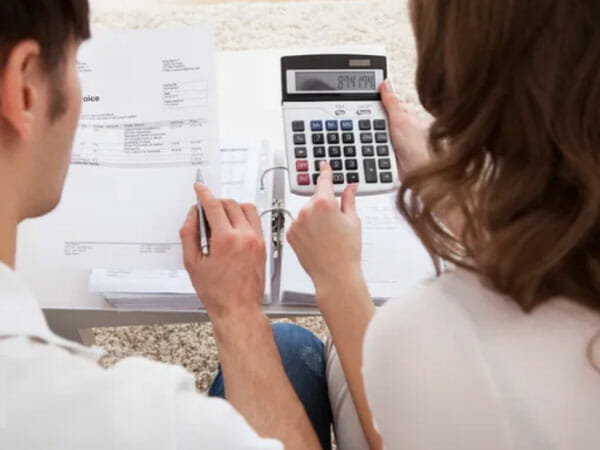 Two people sit at a table reviewing documents. One person uses a calculator while the other holds a pen and looks at papers, possibly calculating finances or paying bills.