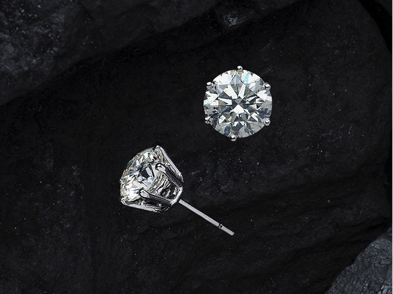 Two round diamond stud earrings with silver prongs are displayed on a dark, textured stone surface.