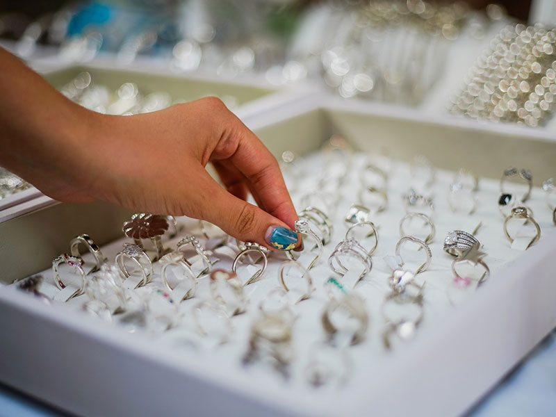 A hand selects a ring from a display case filled with various rings, each arranged in neat rows on a white padded surface.