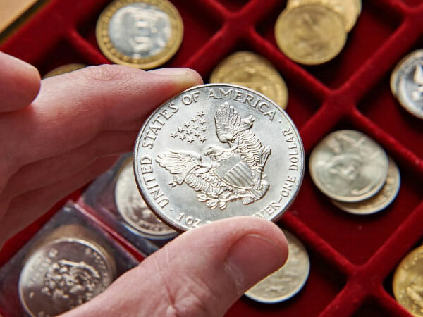A person holds a silver coin with an eagle and “United States of America” text, above a red display case containing various coins.