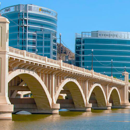 A concrete arch bridge spans over water with modern glass office buildings in the background under a clear blue sky.