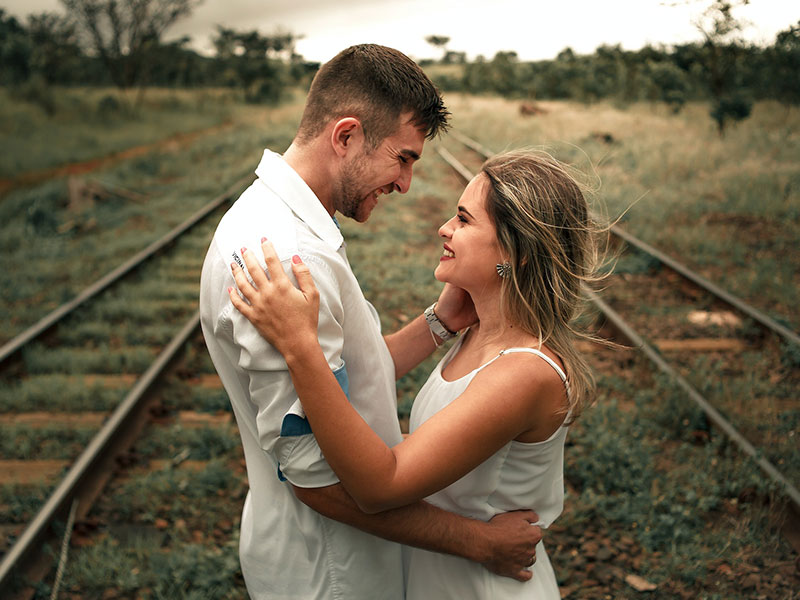 A couple stands facing each other, smiling and embracing on a railway track surrounded by greenery.
